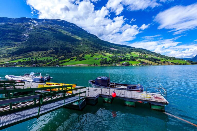 A scenic view of a dock on a tranquil lake, with small boats and vibrant green hills under a partly cloudy sky.