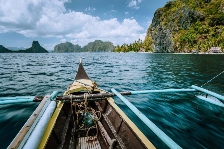 View from a boat's bow navigating through clear blue waters with rocky islands and lush greenery under a partly cloudy sky.