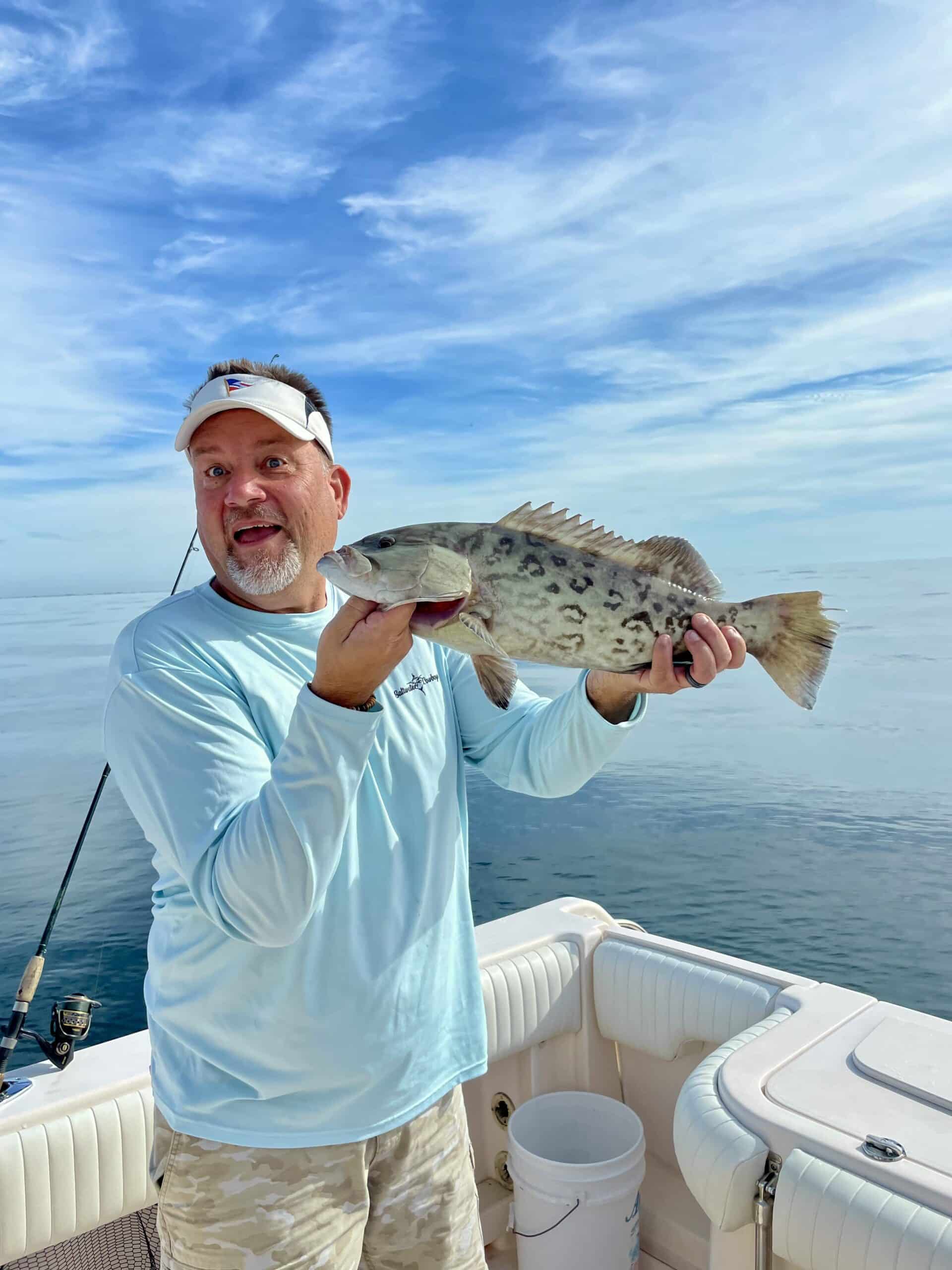 A man on a boat holding a fish with the ocean in the background. He's wearing a light blue shirt, camouflage shorts, and a visor.