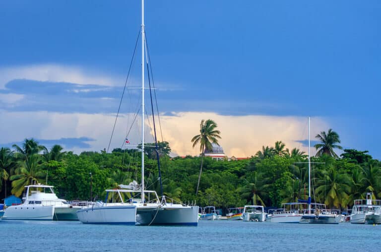 Boats anchored in a calm bay with a backdrop of lush green trees and palm trees under a cloudy sky.