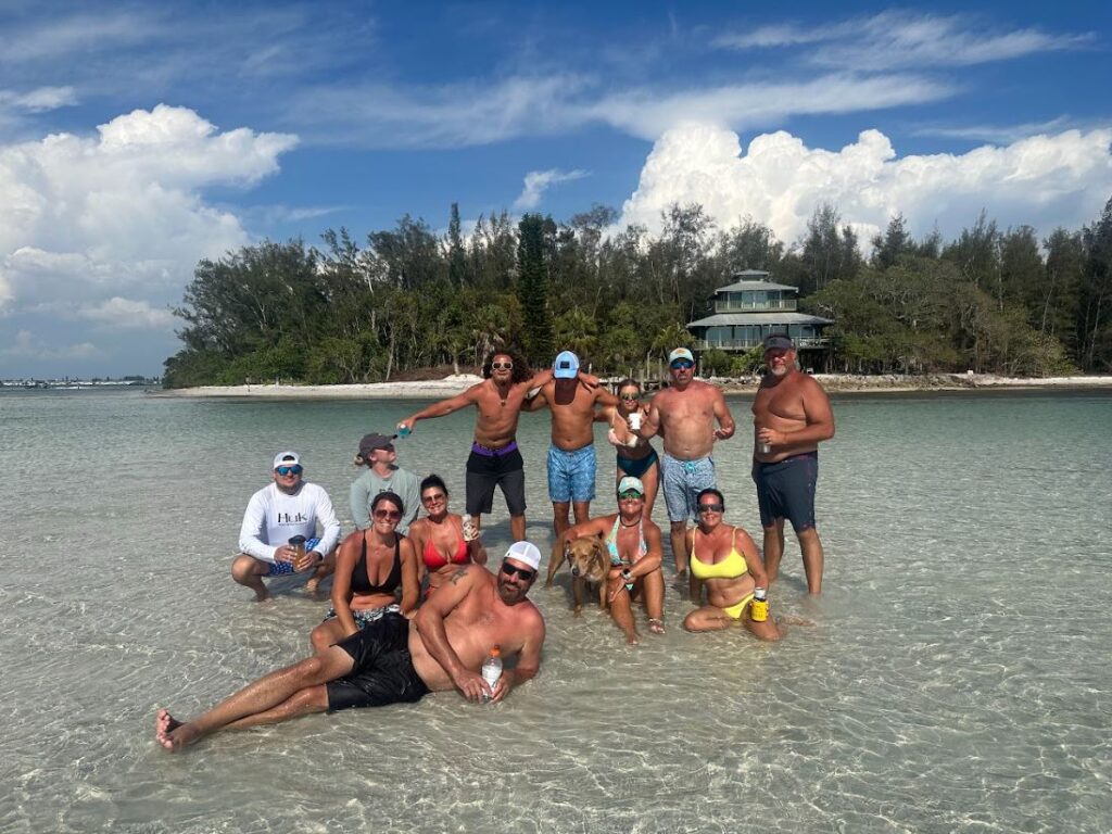 A group of people standing and sitting in shallow water at a beach, with trees and a house in the background under a partly cloudy sky.