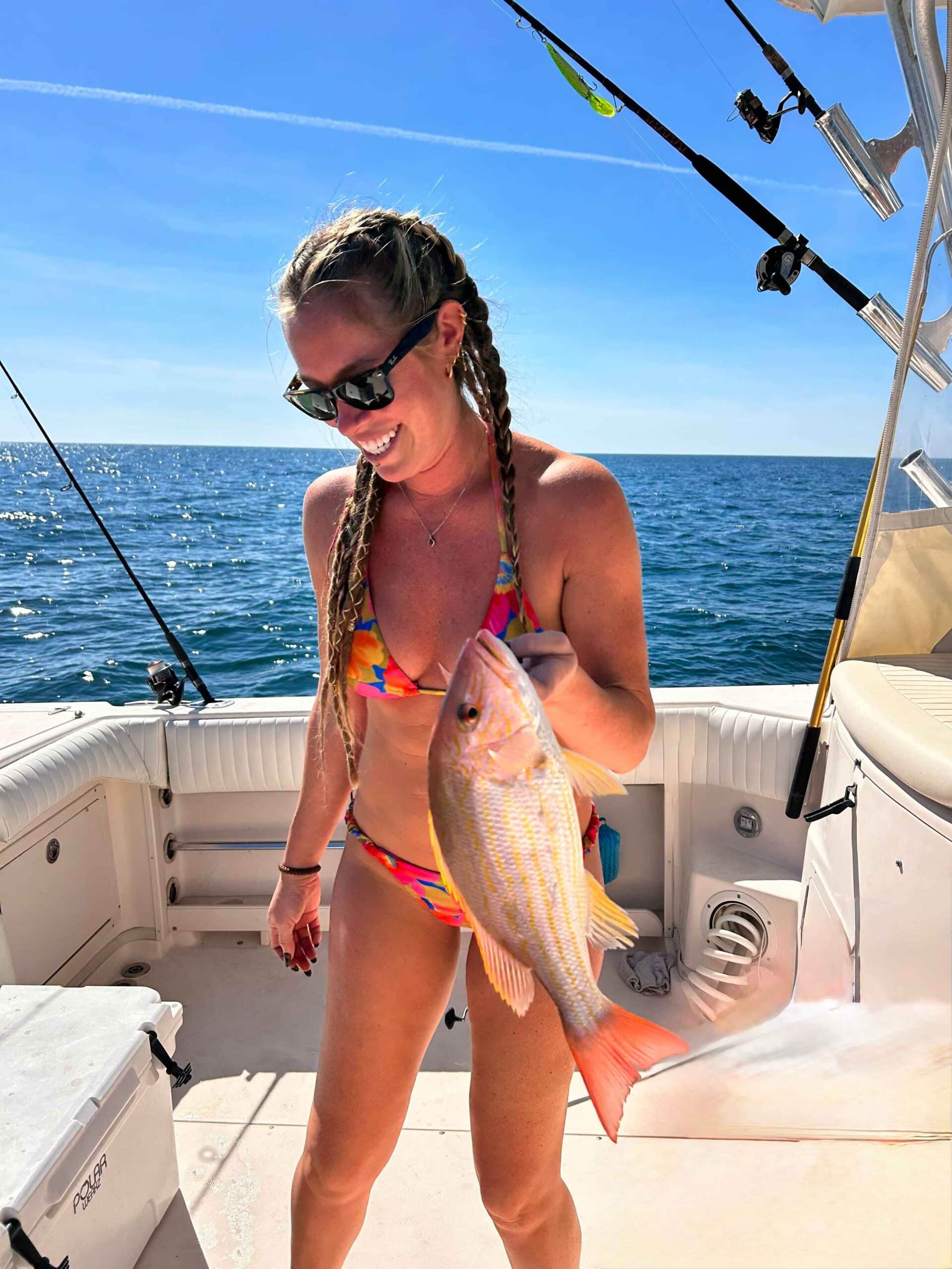 Person in a colorful swimsuit holding a fish on a boat, with fishing rods and the ocean in the background.