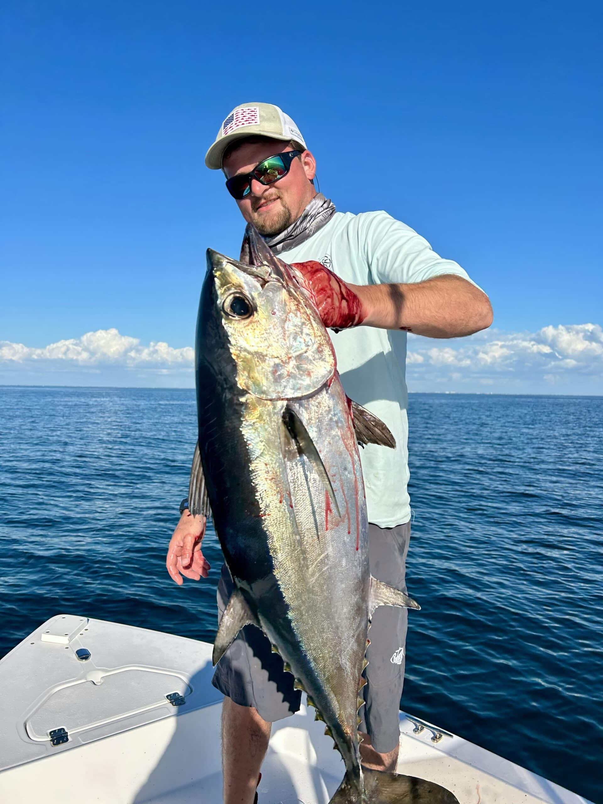 Person holding a large tuna on a boat in open water, wearing a light-colored shirt, hat, sunglasses, and gloves, with a clear blue sky above.