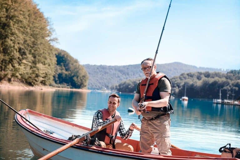 Two men fishing on a small boat in a lake, surrounded by trees and hills. They wear life jackets; one man holds a fishing rod while the other rows. The sky is clear and sunny.