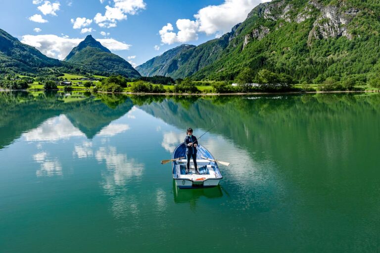 Person standing in a small boat on a calm lake, surrounded by green mountains and a blue sky with clouds.