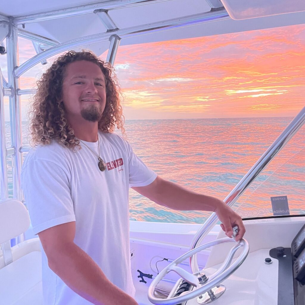 A man with curly hair stands at the helm of a boat, steering, with a vibrant sunset and calm ocean in the background.