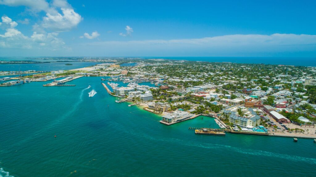 Aerial view of a coastal city with turquoise water, boats, docks, and buildings extending inland under a clear, sunny sky.