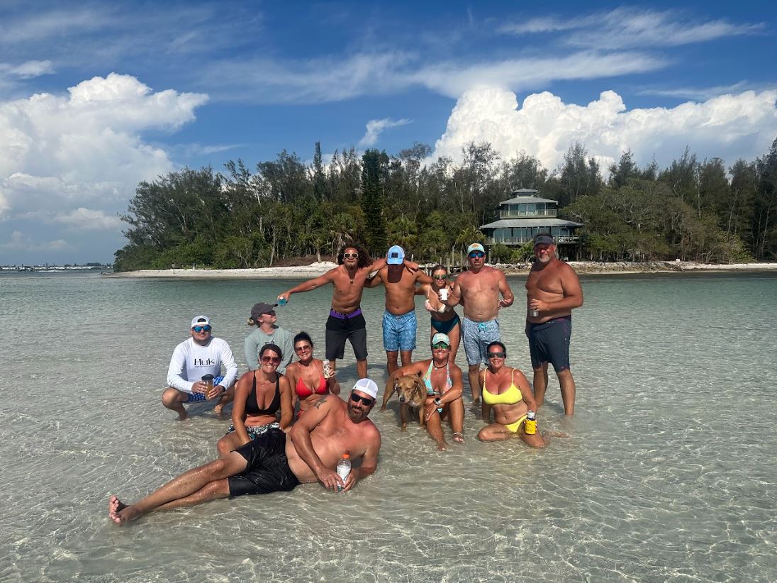 A group of people in swimsuits pose together in shallow, clear water near a sandy beach with trees and a house in the background on a sunny day.