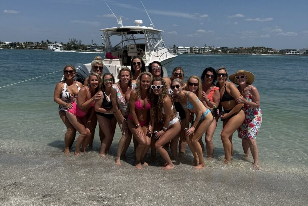 A group of women in swimsuits pose and smile on a beach with shallow water, with a docked boat and waterfront houses visible in the background.