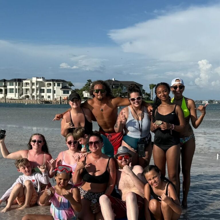 A group of people in swimwear pose and smile together on a sandy beach with buildings and water in the background.