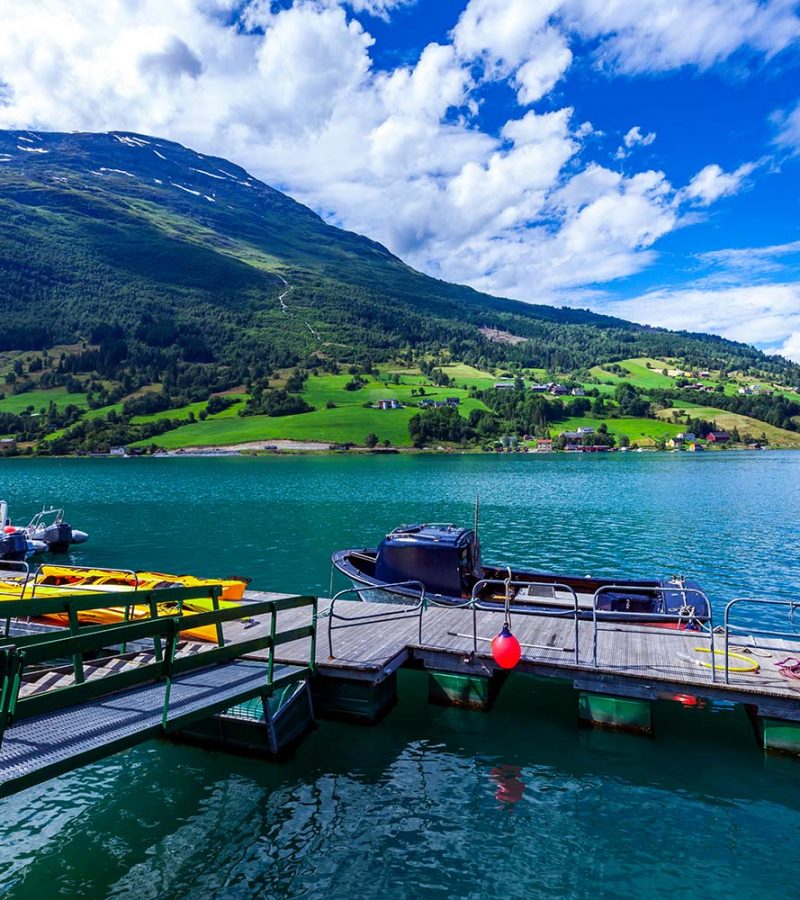 A scenic view of a dock on a tranquil lake, with small boats and vibrant green hills under a partly cloudy sky.