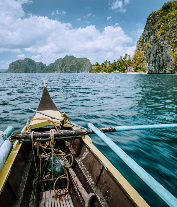 View from a boat's bow navigating through clear blue waters with rocky islands and lush greenery under a partly cloudy sky.