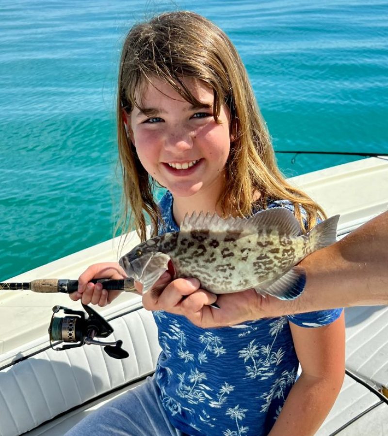 A girl with long hair sits on a boat, smiling and holding a fishing rod. An adult's hand holds a small fish in front of her. The water in the background is calm and blue.