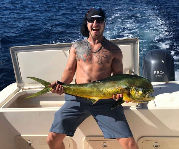 Man sitting on a boat holding a large fish, wearing sunglasses and a hat. The ocean is visible in the background with blue skies above.