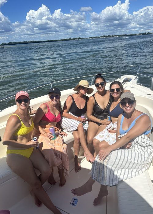 Six women sit together on a boat in swimsuits