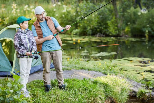 An adult and a child are fishing near a pond, standing on grass. A tent is set up nearby. Both hold fishing rods, focused on the water.