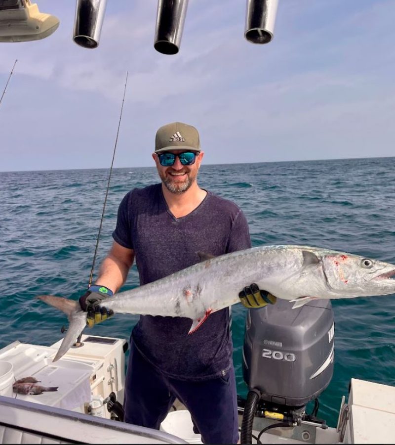 A person on a boat holds a large fish, smiling at the camera. The ocean is visible in the background, along with fishing rods and a motor.
