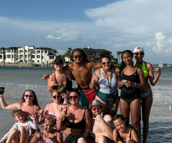 A group of people in swimwear pose and smile together on a sandy beach with buildings and water in the background.