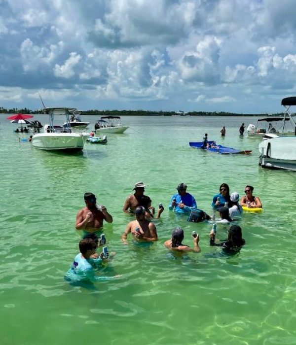 People are gathered in shallow green water, holding drinks. Boats float nearby under a partly cloudy sky.