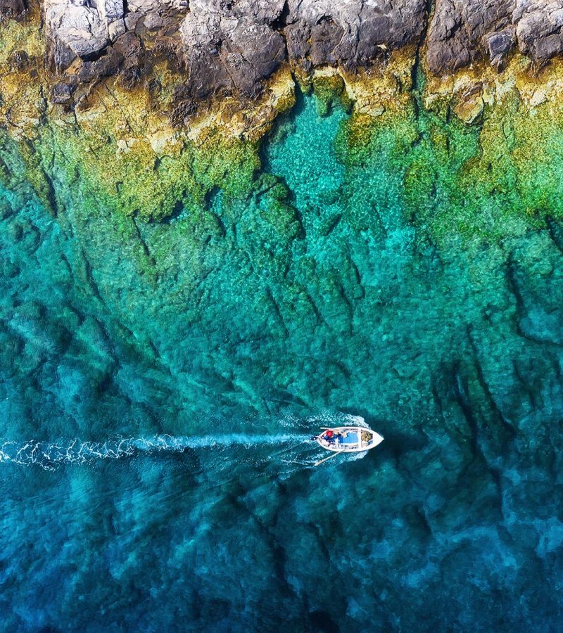 A small boat travels across clear turquoise waters near a rocky shoreline, leaving a visible wake behind.