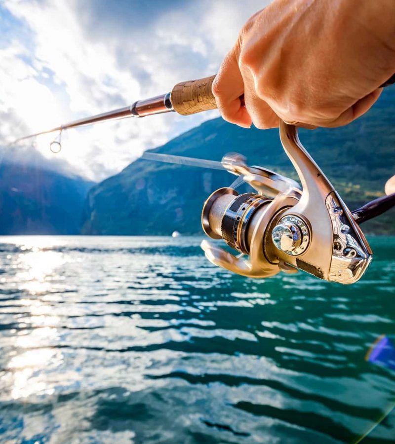 Person fishing with a rod and reel by a mountain lake under a partly cloudy sky, sunlight reflecting on the water.